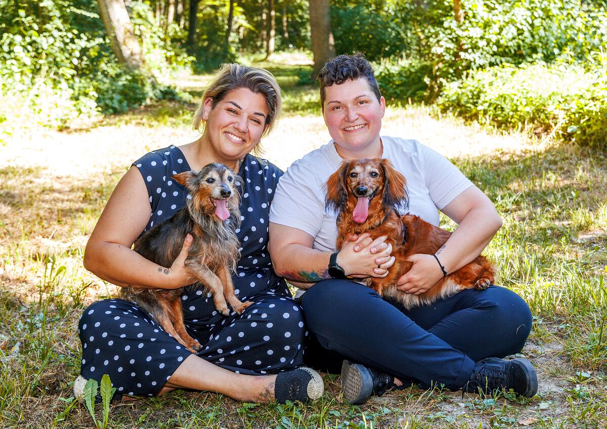 Paula (links) und Michaela (rechts) sitzen mit ihren beiden Hunden auf einer sonnigen Waldlichtung